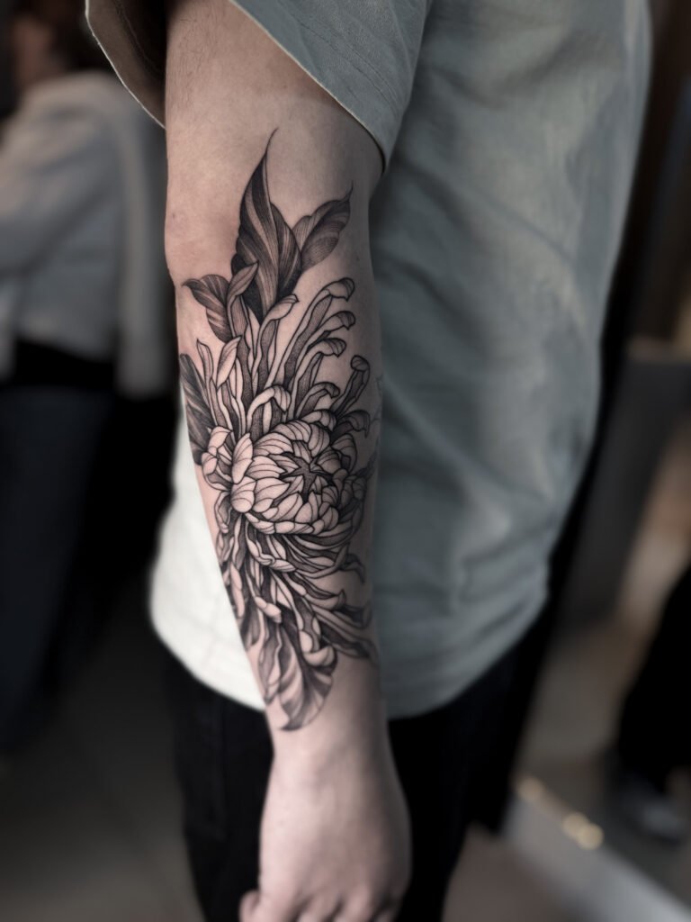 Black and grey realistic tattoo of a chrysanthemum flower on the forearm, symbolizing rebirth and strength. Detailed shading and depth by NOIR Tattoo Cusco, Peru.