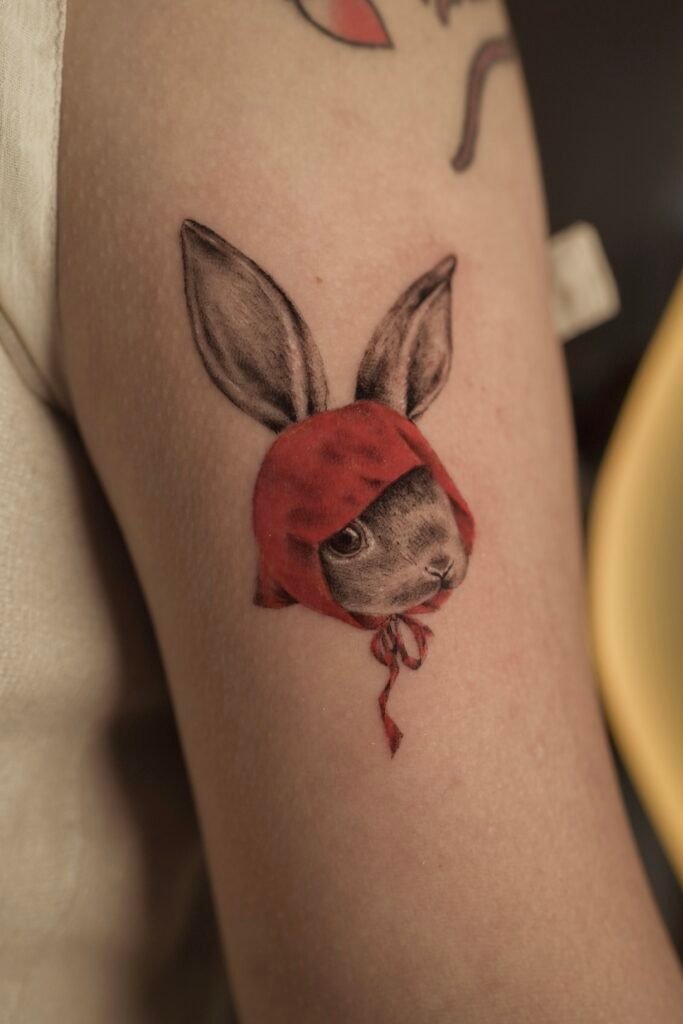 Realistic tattoo of a bunny wearing a red hood, blending innocence and surreal symbolism. Created at NOIR Tattoo Cusco, Peru.