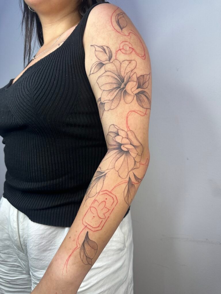Sketch-style floral sleeve tattoo with red ink details on the arm, featuring elegant linework and minimalist botanical design. Created at NOIR Tattoo Cusco, Peru.