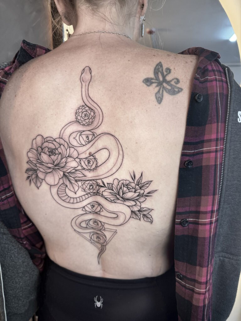 Black and grey back tattoo featuring a coiled snake intertwined with peonies and lotus flowers, symbolizing transformation, renewal, and feminine energy — created at NOIR Tattoo Cusco, Peru.