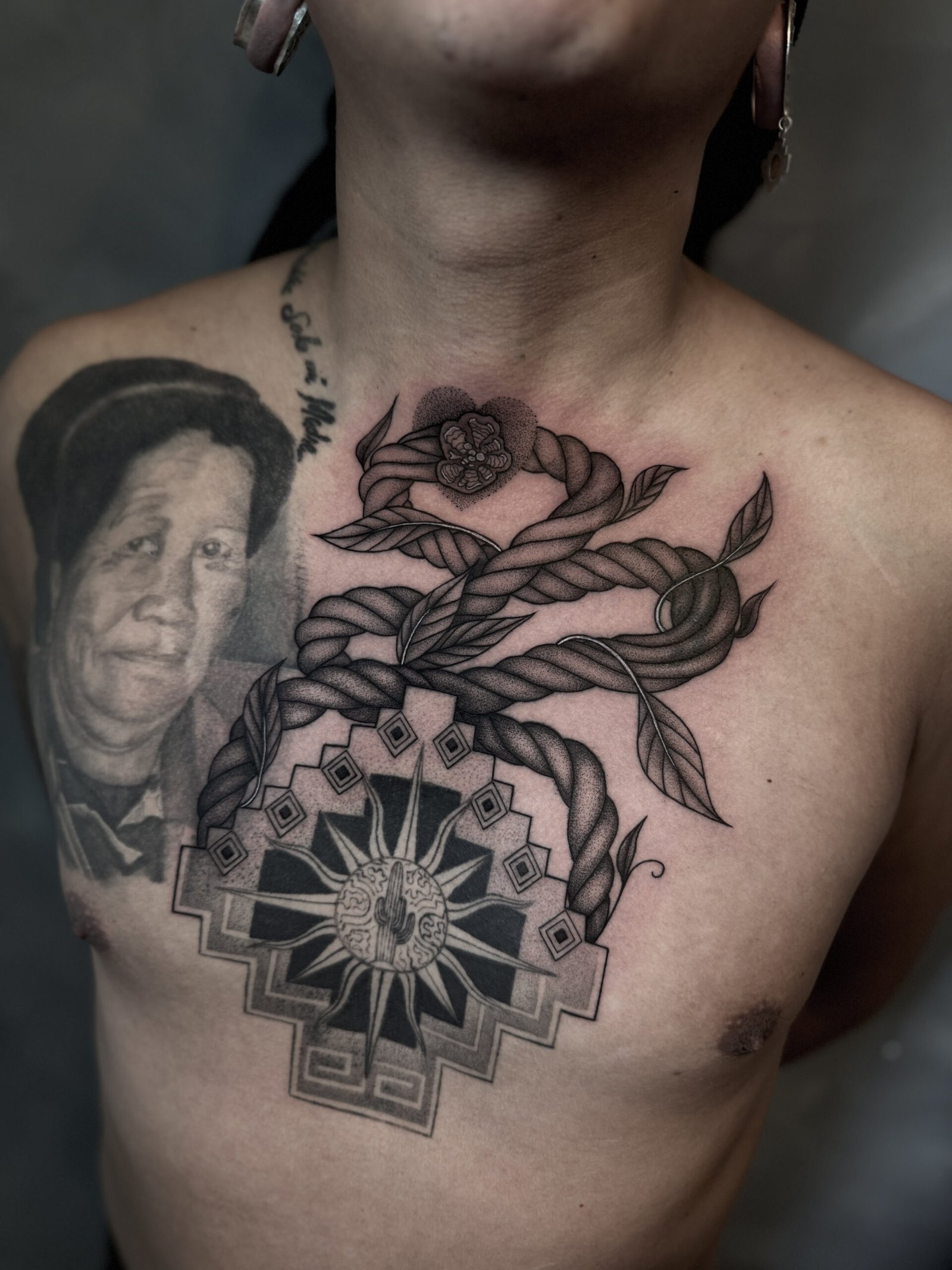 Black and grey chest tattoo of an ayahuasca vine (Banisteriopsis caapi) with leaves and flower intertwined with an Andean geometric sun/mandala, symbolizing healing, vision, and protection — created at NOIR Tattoo Cusco, Peru.