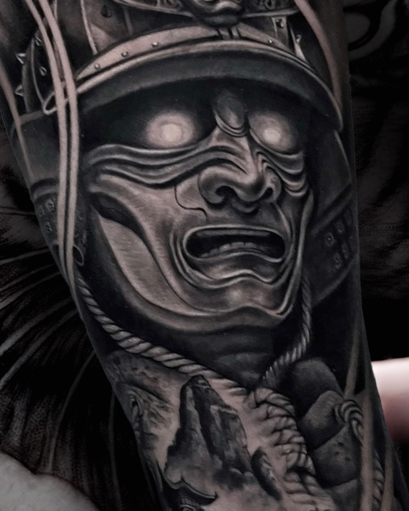 Black and grey realism tattoo of a samurai warrior mask with glowing eyes and armor details, symbolizing honor, discipline, and strength, created at NOIR Tattoo Studio in Cusco, Peru.