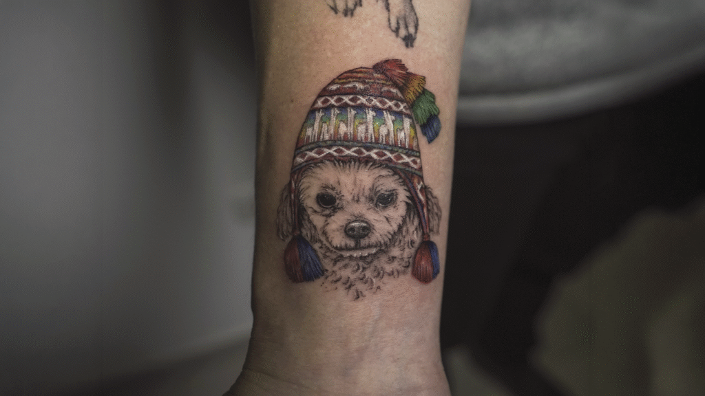 Cute dog portrait tattoo wearing a traditional Andean chullo hat, created in black and grey with colorful details at Noir Tattoo Cusco, Peru.