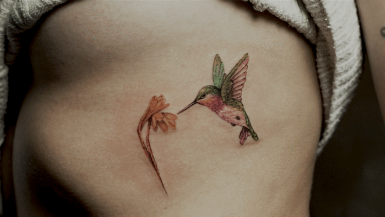 Delicate fine line hummingbird tattoo with a flower in soft watercolor colors, tattooed at Noir Tattoo Cusco, Peru.
