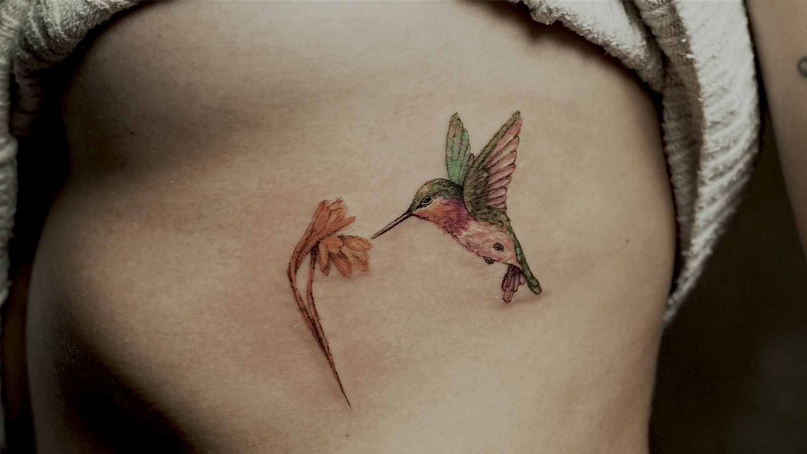 Delicate fine line hummingbird tattoo with a flower in soft watercolor colors, tattooed at Noir Tattoo Cusco, Peru.