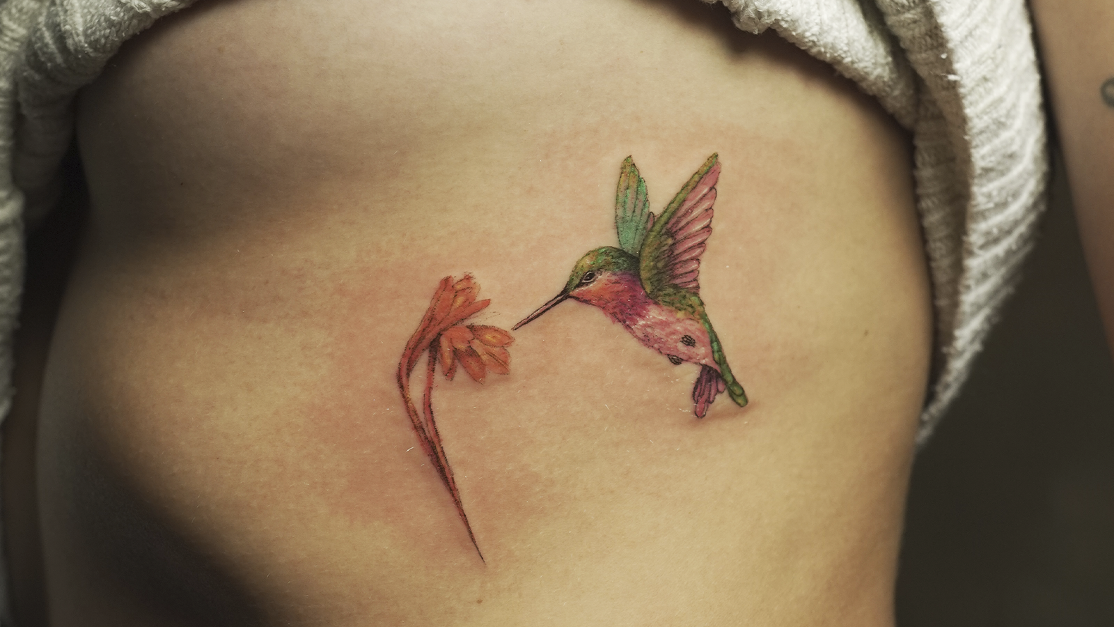 Color hummingbird tattoo hovering beside a flower on the rib cage, created at Noir Tattoo Cusco, Peru.