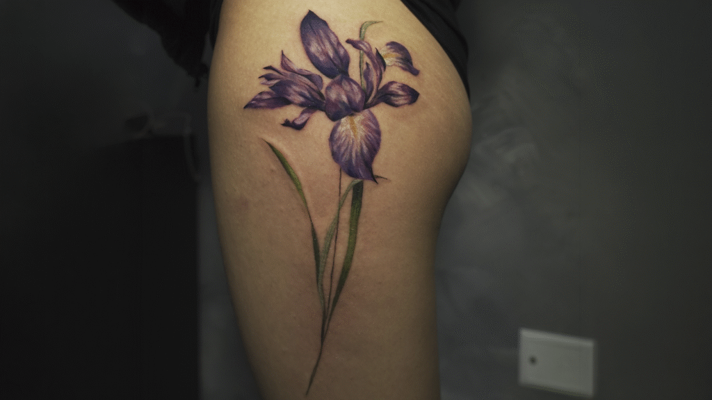 Purple iris flower tattoo on the thigh featuring realistic color shading and botanical details, created at Noir Tattoo Cusco.
