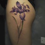 Purple iris flower tattoo on the thigh featuring realistic color shading and botanical details, created at Noir Tattoo Cusco.