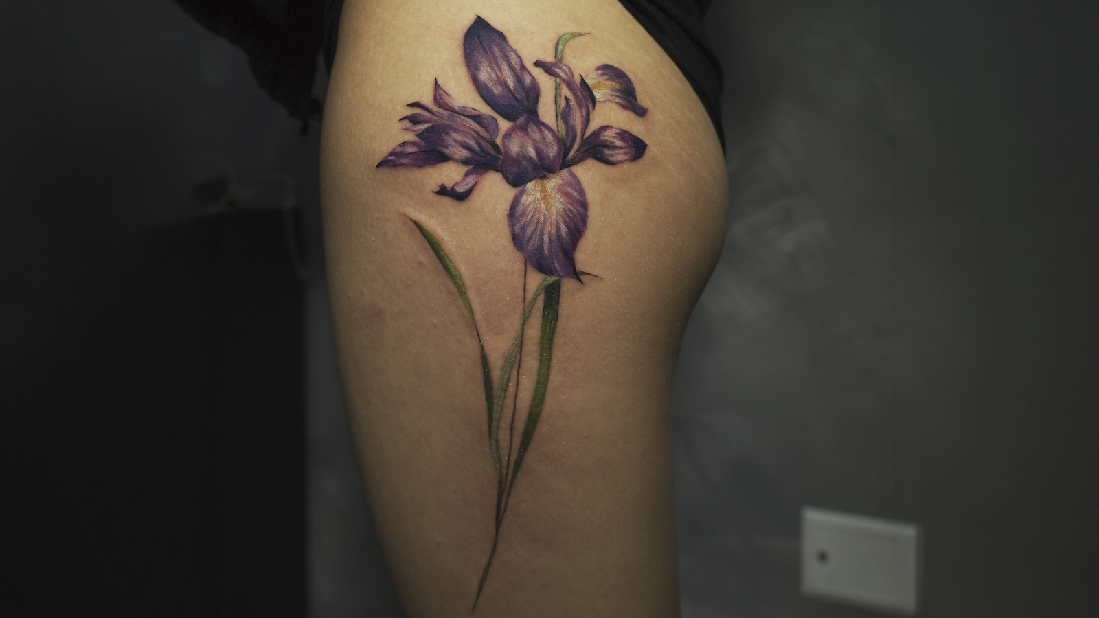 Purple iris flower tattoo on the thigh featuring realistic color shading and botanical details, created at Noir Tattoo Cusco.